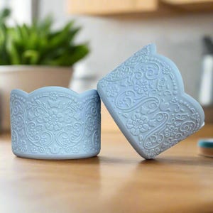 May include: Two light blue silicone cupcake liners with a delicate floral pattern. The liners are made of a flexible material and are designed to be reusable.