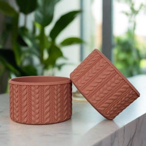 May include: Two terracotta colored ceramic planters with a cable knit pattern. The planters are round and have a smooth finish.