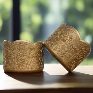 May include: Two gold-colored metal cupcake liners with a decorative floral pattern.