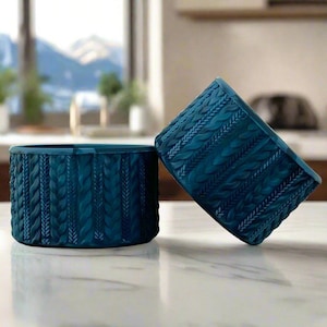 May include: Two teal blue ceramic bowls with a knitted pattern. The bowls are on a white marble surface.