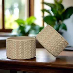 May include: Two beige ceramic candle holders with a knitted pattern. The holders are cylindrical and have a smooth, matte finish.
