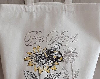 May include: A white canvas tote bag with black handles. The bag has a black and yellow bee and flowers printed on it. The text "Be Kind" is printed in cursive above the bee and flowers.