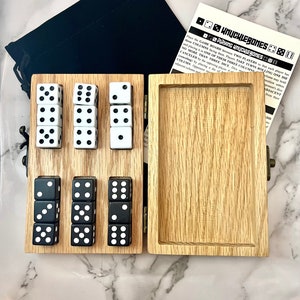 May include: A wooden box with a hinged lid, containing a set of black and white dice. The box is open, revealing the dice inside. The box is labelled "Knuckle Bones Playing Knuckle Bones".