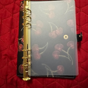 May include: Black planner with gold-colored rings and a clear cover. The planner's interior features a cherry pattern. The cover is closed with a black snap. The planner is on a red quilted surface.