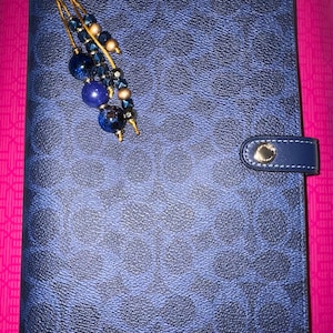 May include: A rectangular blue planner cover featuring a dark blue repeating logo pattern. A gold-toned metal snap closure secures the side. A decorative charm hangs from the top, consisting of blue and metallic beads on gold-toned wire strands.