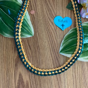 May include: A braided necklace in teal and gold. The necklace is made of woven ribbon, with each section alternating between teal and gold. The necklace is displayed on a wooden surface with green leaves and a blue heart-shaped tag.
