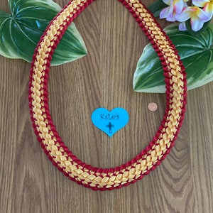 May include: A braided lei necklace with alternating gold and red ribbons. The lei is arranged in a circular shape, with a red and gold ribbon bow at the top. A blue heart with the text "KsLeis" and a cross is visible.
