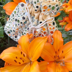 May include: A white fabric pouch with a drawstring closure decorated with small round mirrors. The pouch is lying on top of two orange lilies.