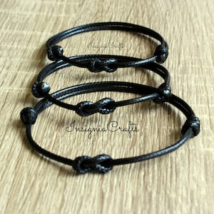 May include: Three black friendship bracelets with infinity knots. The bracelets are made of a thin, braided cord and are stacked on top of each other. The text "Insignia Crafts" is visible on the bottom bracelet.