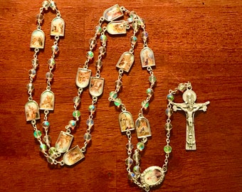 May include: A silver rosary with clear glass beads and silver medallions featuring images of saints. The rosary has a silver crucifix with a figure of Jesus Christ.