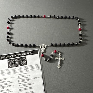 May include: Black rosary beads with red accents and a silver crucifix. The beads are strung on a silver chain. The rosary is accompanied by a printed card with the text "The Chaplet for the Dead" and a prayer.