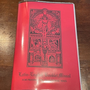 May include: A red booklet titled "Latin-English Booklet Missal" with a detailed black illustration of a religious scene. The booklet is encased in a clear plastic cover and is designed for praying the traditional mass.