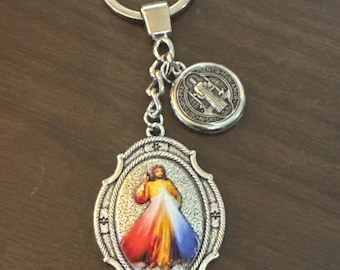 May include: A silver-toned metal keychain featuring an oval pendant with a textured border and a colorful image of Jesus Christ. A smaller, round Saint Benedict medal is attached to the chain, which connects to a circular split ring.
