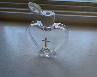 Puede incluir: Una botella de plástico transparente con forma de corazón con una tapa blanca y una cruz dorada y las palabras "Holy Water" impresas en la parte delantera.