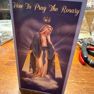 May include: A religious book titled "How To Pray The Rosary" features an illustration of a woman in a blue and white robe, standing on clouds. The image has a purple background with gold accents and a gold crown. The book is displayed on a wooden surface.