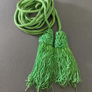 Puede incluir: Un par de cordones con borlas verdes con un cordón verde largo. Las borlas están hechas de tejido tejido y tienen flecos.