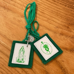 May include: Two green fabric prayer cards with white trim and white text. One card features an image of Saint Bernadette and the text "Immaculate Heart of Mary Pray For Us". The other card features a heart with a cross and the text "Now and at the hour of our death". A green cord is attached to the cards.