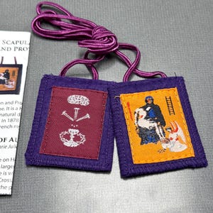 May include: Two purple fabric squares with embroidered images. The left square has a white image of a chalice with three nails above it. The right square has a yellow background with a white image of a man in a black robe holding a white cross. The squares are connected by a purple cord.