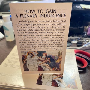 May include: A white card with brown text explaining how to gain a plenary indulgence. The card features a painting of Jesus being carried by two men, with a woman in a blue robe and a man in a turban behind them. The painting is set against a backdrop of a city with buildings and trees.