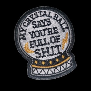 Peut inclure: Un patch brodé blanc en forme de boule de cristal avec le texte "My Crystal Ball Says You're Full Of Shit" en noir avec un éclair et une lune croissante dorés.