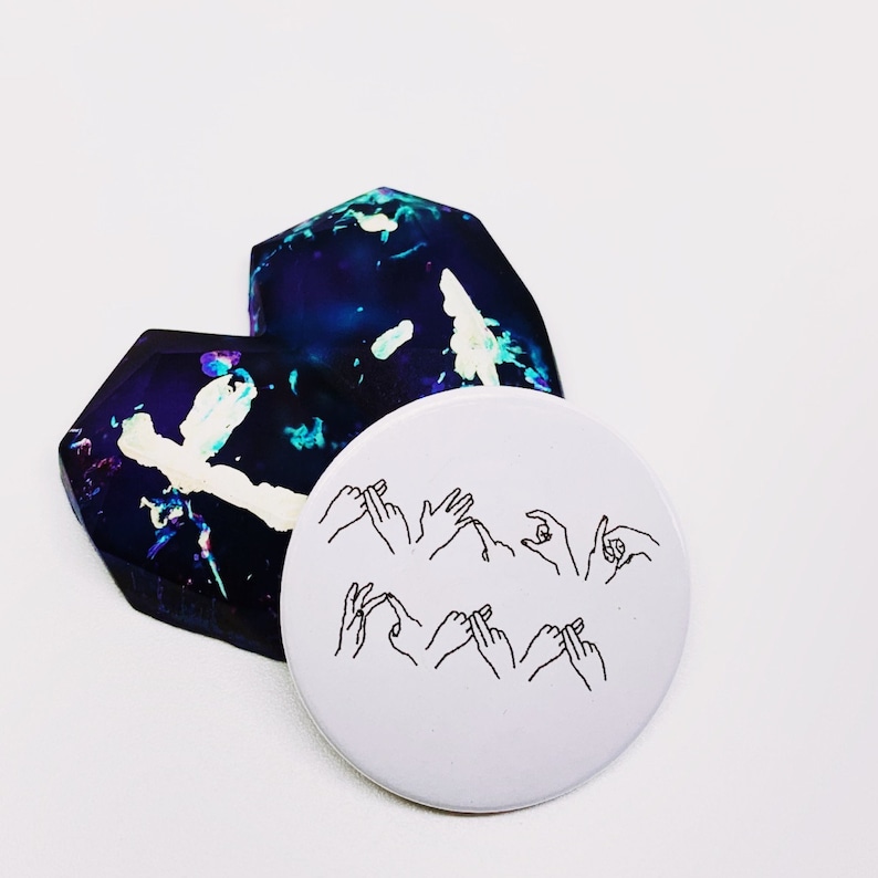 May include: A white round sticker with black line drawings of hands signing the word "love" in American Sign Language. The sticker is on top of a heart-shaped piece of iridescent glass.