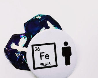 May include: A white round button with black text and a black stick figure. The text on the button reads "26 Fe 55.845". The button is sitting on top of two blue and purple resin coasters.