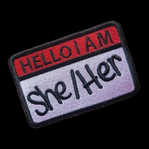 Op de afbeelding: Een rood en zwart naambordje met de tekst "HELLO I AM" in rood en "She/Her" in zwart.