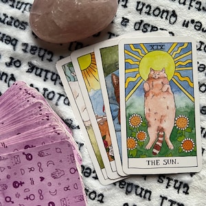 May include: A tarot card reading with a pink crystal, a deck of tarot cards with purple symbols, and three tarot cards showing the sun, a cat, and a landscape. The top card shows a cat with a sun behind it and the text "THE SUN."