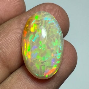 May include: An oval-shaped opal gemstone with a mosaic-like pattern. The opal displays a play of color, with flashes of green, yellow, orange, and blue. The stone has a polished, glossy surface, and is held between fingers.