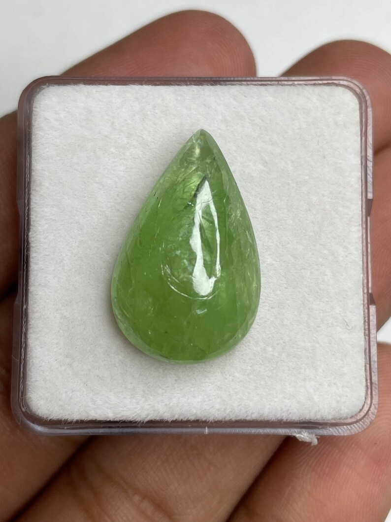 Puede incluir: Una gema verde claro en forma de pera, exhibida en una caja de pl&aacute;stico transparente con un interior blanco texturizado. La gema pulida tiene una superficie lisa y un aspecto transl&uacute;cido, mostrando su color natural y caracter&iacute;sticas internas.