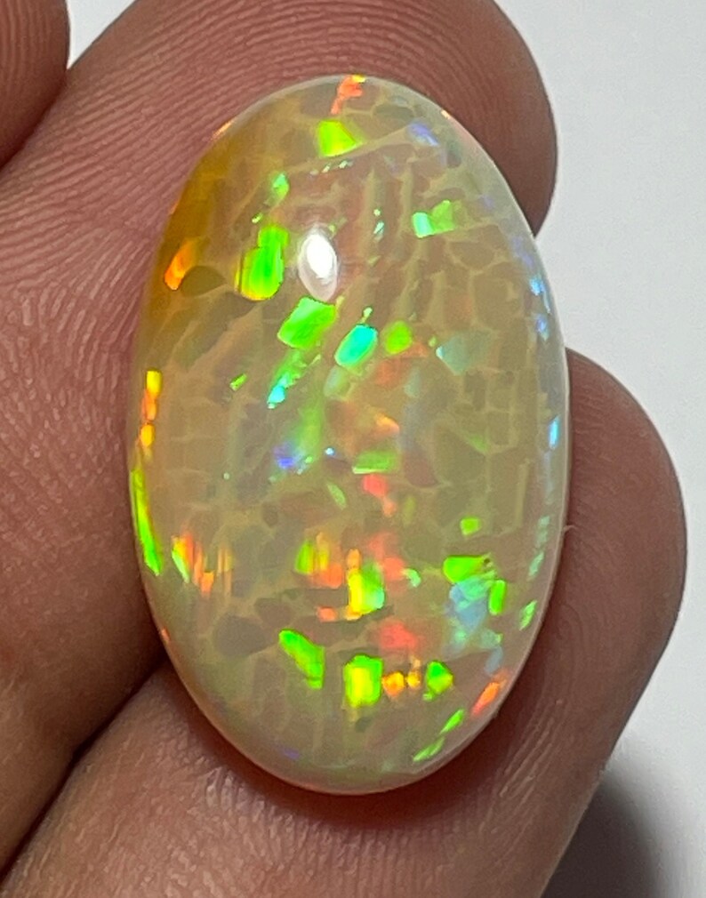 May include: Oval-shaped opal gemstone with a cream-colored base and vibrant flashes of green, orange, and yellow. The gemstone has a unique internal pattern and is held in a person's hand. The opal is a natural gemstone.
