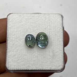 K&ouml;nnte beinhalten: Zwei ovale Edelsteine mit blauen und gr&uuml;nen Farbt&ouml;nen werden in einer transparenten Kunststoffbox mit wei&szlig;em Innenraum pr&auml;sentiert. Die Edelsteine weisen interne Aderungen auf, die ein einzigartiges Aussehen erzeugen. Die Box befindet sich vor einem neutralen Hintergrund.
