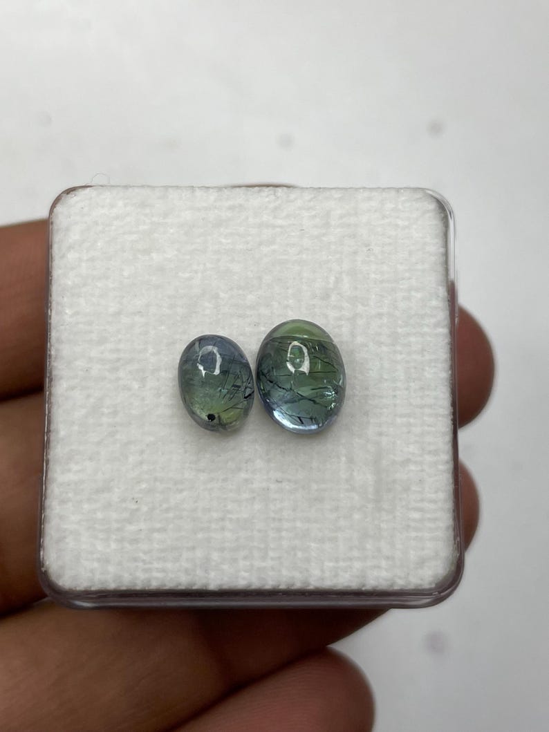 K&ouml;nnte beinhalten: Zwei ovale Edelsteine mit blauen und gr&uuml;nen Farbt&ouml;nen werden in einer transparenten Kunststoffbox mit wei&szlig;em Innenraum pr&auml;sentiert. Die Edelsteine weisen interne Aderungen auf, die ein einzigartiges Aussehen erzeugen. Die Box befindet sich vor einem neutralen Hintergrund.