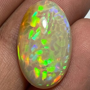 May include: Oval-shaped opal gemstone with a vibrant display of color. The stone features flashes of green, orange, and blue hues within a translucent, milky white base. The opal's surface is polished, showcasing its internal play of color.