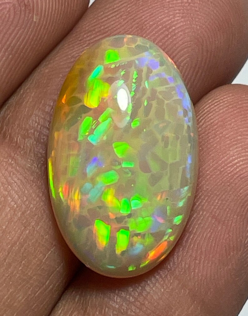 May include: Oval-shaped opal gemstone with a vibrant display of color. The stone features flashes of green, orange, and blue hues within a translucent, milky white base. The opal's surface is polished, showcasing its internal play of color.