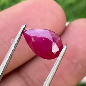 May include: A faceted, pear-shaped ruby gemstone held by tweezers. The ruby is a deep, vibrant red colour, with multiple facets catching the light. The gemstone is displayed against a blurred green background.