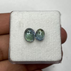 K&ouml;nnte beinhalten: Zwei ovale Edelsteine mit blau-gr&uuml;nen Farbt&ouml;nen werden in einer transparenten Kunststoffbox mit wei&szlig;em Innenraum pr&auml;sentiert. Die Edelsteine sind vor einem wei&szlig;en Hintergrund platziert, um ihre Farbe und Klarheit hervorzuheben.