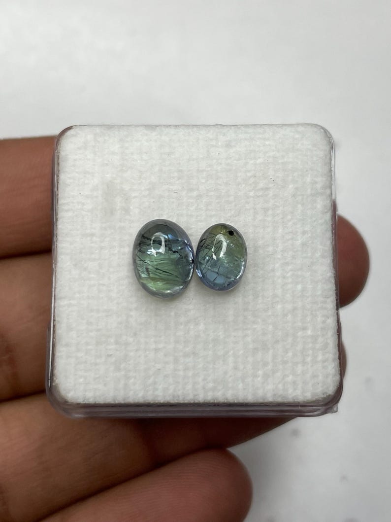 K&ouml;nnte beinhalten: Zwei ovale Edelsteine mit blau-gr&uuml;nen Farbt&ouml;nen werden in einer transparenten Kunststoffbox mit wei&szlig;em Innenraum pr&auml;sentiert. Die Edelsteine sind vor einem wei&szlig;en Hintergrund platziert, um ihre Farbe und Klarheit hervorzuheben.