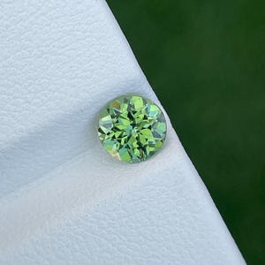 Op de afbeelding: Een ronde, gefacetteerde edelsteen met een levendige limoengroene kleur. De steen heeft een briljante slijpvorm met talloze driehoekige en vliegervormige facetten die het licht reflecteren, wat zorgt voor een sprankelend uiterlijk.
