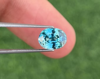 May include: An oval-shaped, light blue gemstone is held by tweezers against a blurred green background. The gemstone has a faceted surface, reflecting light. The gem's colour is a vibrant, clear blue. The gem is likely a precious stone.