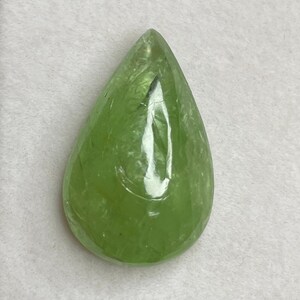 Puede incluir: Una gema transl&uacute;cida en forma de pera, con un tono verde claro vibrante. La superficie pulida refleja la luz, mostrando la claridad y la estructura interna de la piedra. La gema se presenta sobre un fondo neutro, resaltando su belleza natural.