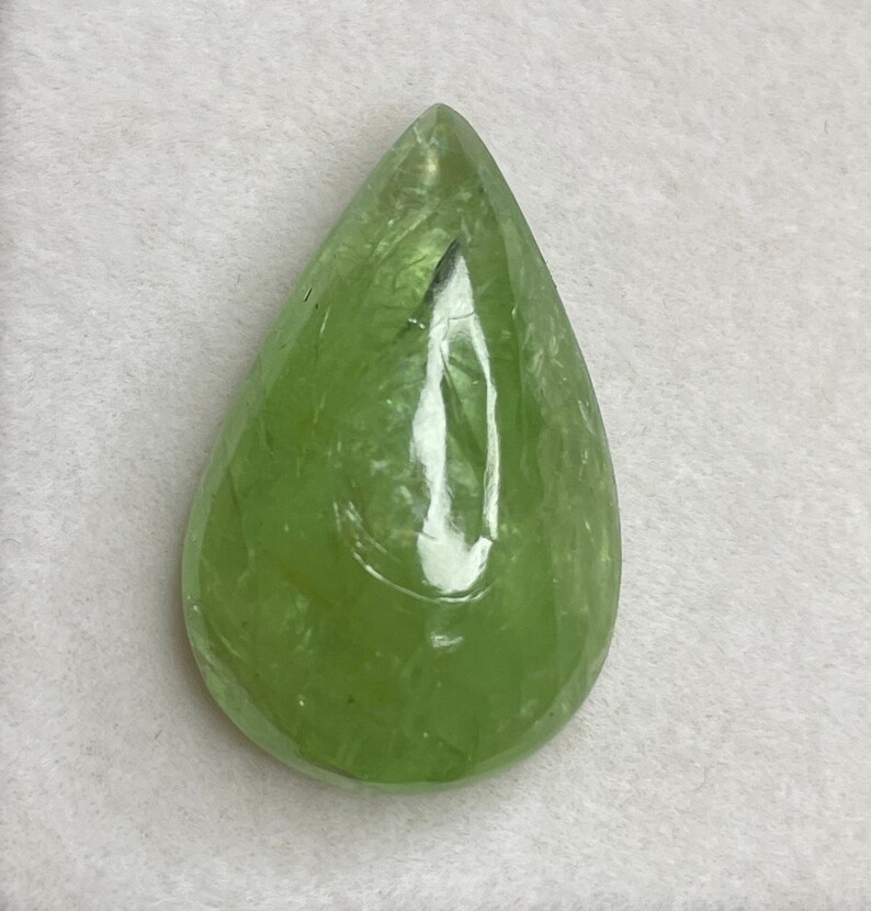 Puede incluir: Una gema transl&uacute;cida en forma de pera, con un tono verde claro vibrante. La superficie pulida refleja la luz, mostrando la claridad y la estructura interna de la piedra. La gema se presenta sobre un fondo neutro, resaltando su belleza natural.