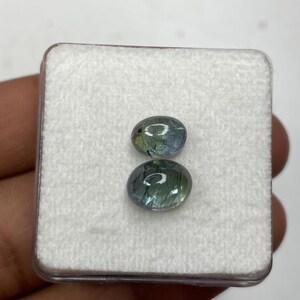K&ouml;nnte beinhalten: Zwei ovale Edelsteine in einem transparenten Kunststoffetui. Die Steine haben eine Mischung aus Blau, Gr&uuml;n und klaren T&ouml;nen. Sie haben eine polierte Oberfl&auml;che und liegen auf einem wei&szlig;en, strukturierten Hintergrund. F&uuml;r Schmuck geeignet.