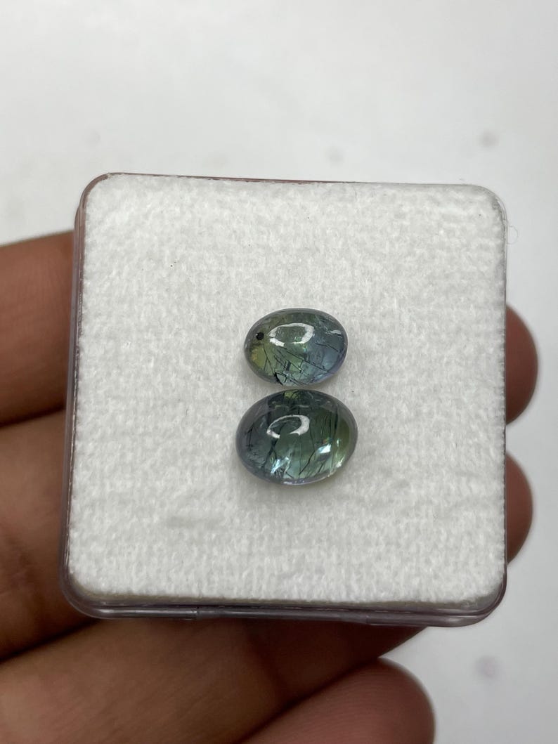 K&ouml;nnte beinhalten: Zwei ovale Edelsteine in einem transparenten Kunststoffetui. Die Steine haben eine Mischung aus Blau, Gr&uuml;n und klaren T&ouml;nen. Sie haben eine polierte Oberfl&auml;che und liegen auf einem wei&szlig;en, strukturierten Hintergrund. F&uuml;r Schmuck geeignet.