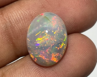 May include: An oval-shaped opal gemstone with a milky white base color. The opal displays vibrant flashes of color, including red, orange, yellow, green, and blue, creating a play-of-color effect. The gemstone is held in a hand.