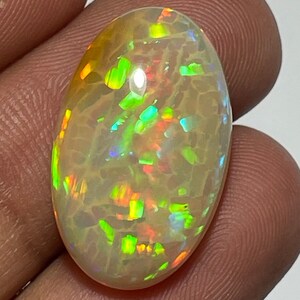 May include: An oval-shaped opal gemstone with a creamy white base color. The opal displays vibrant flashes of green, orange, and blue, creating a play-of-color effect. The stone is held between fingers, showcasing its polished surface.