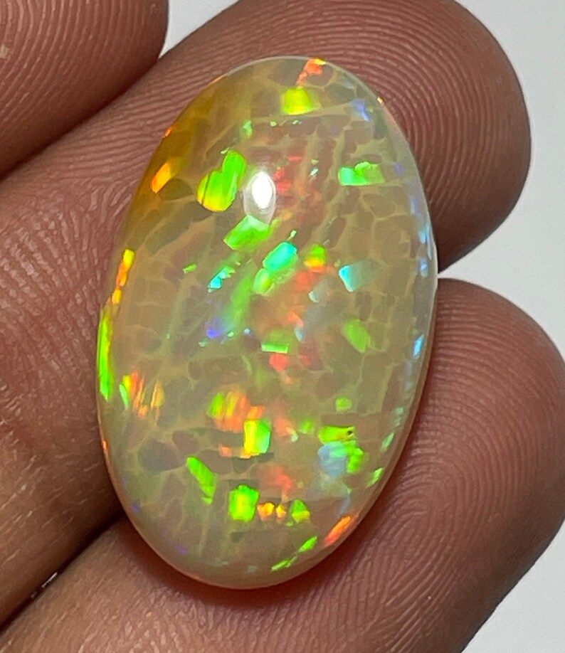 May include: An oval-shaped opal gemstone with a creamy white base color. The opal displays vibrant flashes of green, orange, and blue, creating a play-of-color effect. The stone is held between fingers, showcasing its polished surface.