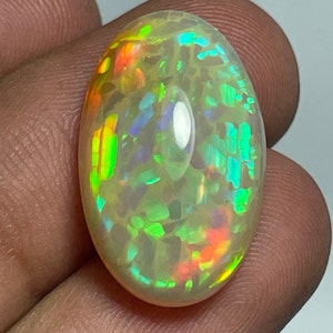 May include: An oval-shaped opal gemstone with a milky white base and vibrant flashes of color, including green, orange, and blue. The gemstone is polished and smooth, reflecting light and showcasing its internal play-of-color.