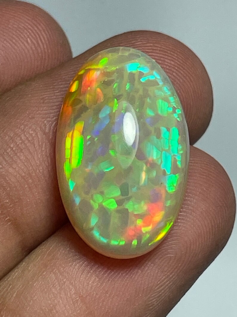 May include: An oval-shaped opal gemstone with a milky white base and vibrant flashes of color, including green, orange, and blue. The gemstone is polished and smooth, reflecting light and showcasing its internal play-of-color.
