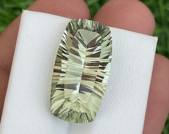 May include: A rectangular, faceted gemstone with a pale green hue. The gem is held against a white surface, showcasing its intricate cuts and internal reflections. The stone's shape is elongated with rounded ends, and it appears to be a polished, high-quality gemstone.